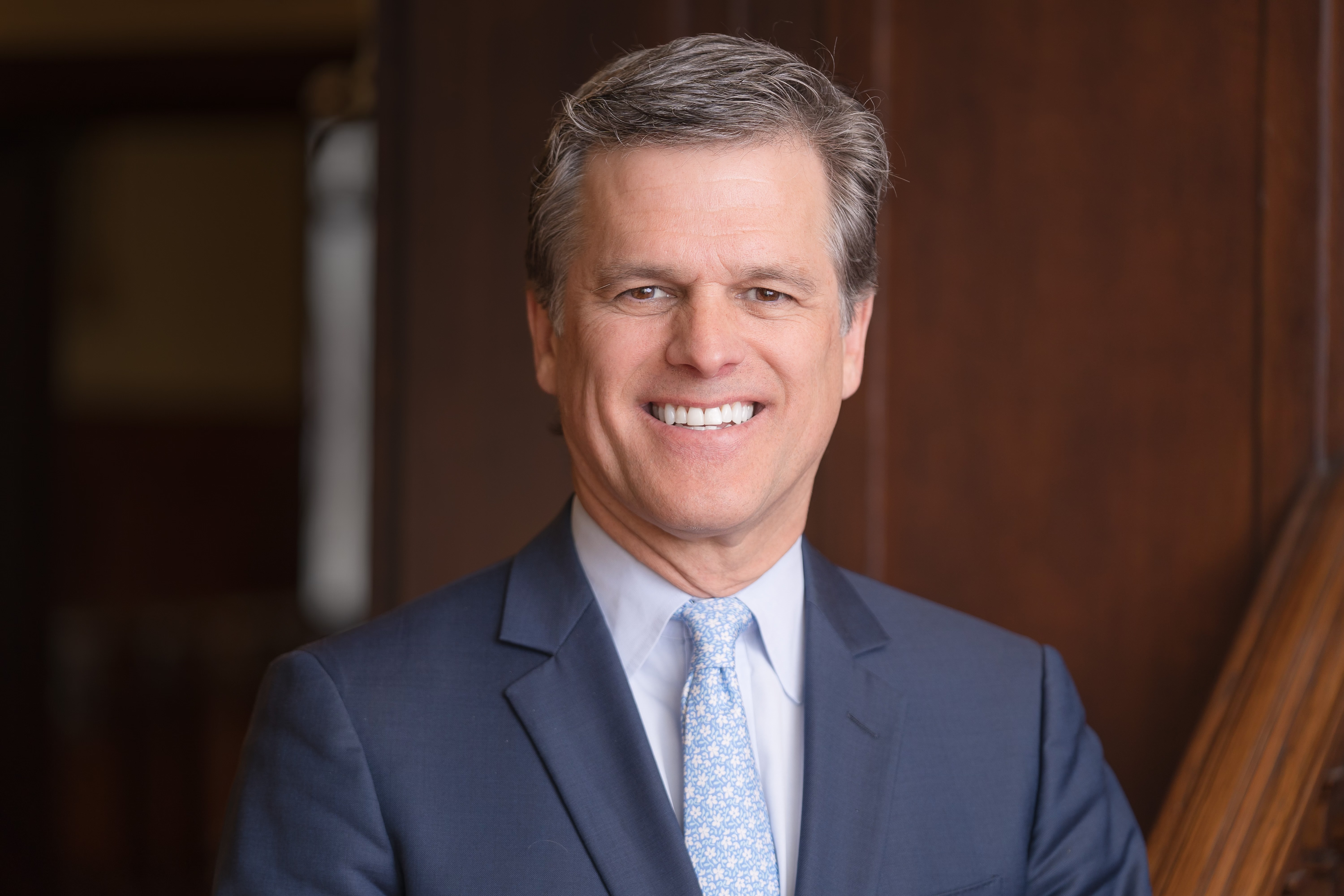 Tim Shriver - light skinned male in a suit smiling with neatly combed gray hair in front of a wood tone background